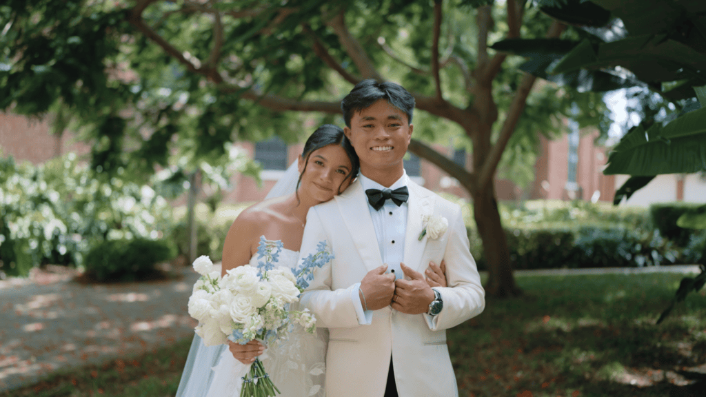 Bride (left) Mary Grace stands next to her husband (right) Miggy holding a bouquet of white and blue flowers. Her head is nuzzled on Miggy's shoulder. Both smile.