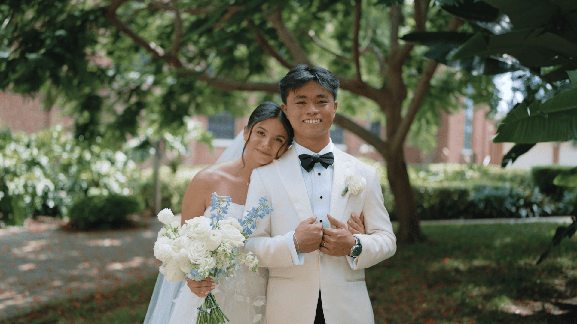 Bride (left) Mary Grace stands next to her husband (right) Miggy holding a bouquet of white and blue flowers. Her head is nuzzled on Miggy's shoulder. Both smile.